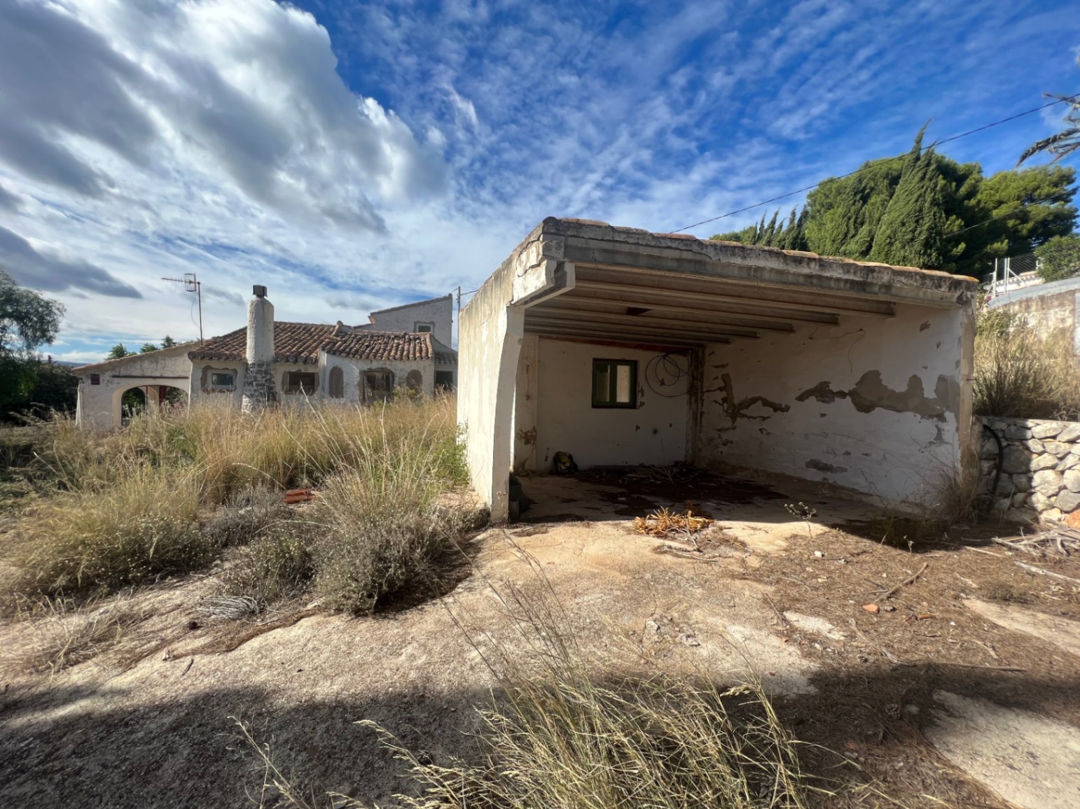 Cochera independiente y trastero en estado original, situados en una amplia parcela con vegetación natural y vistas a la vivienda principal de estilo mediterráneo.