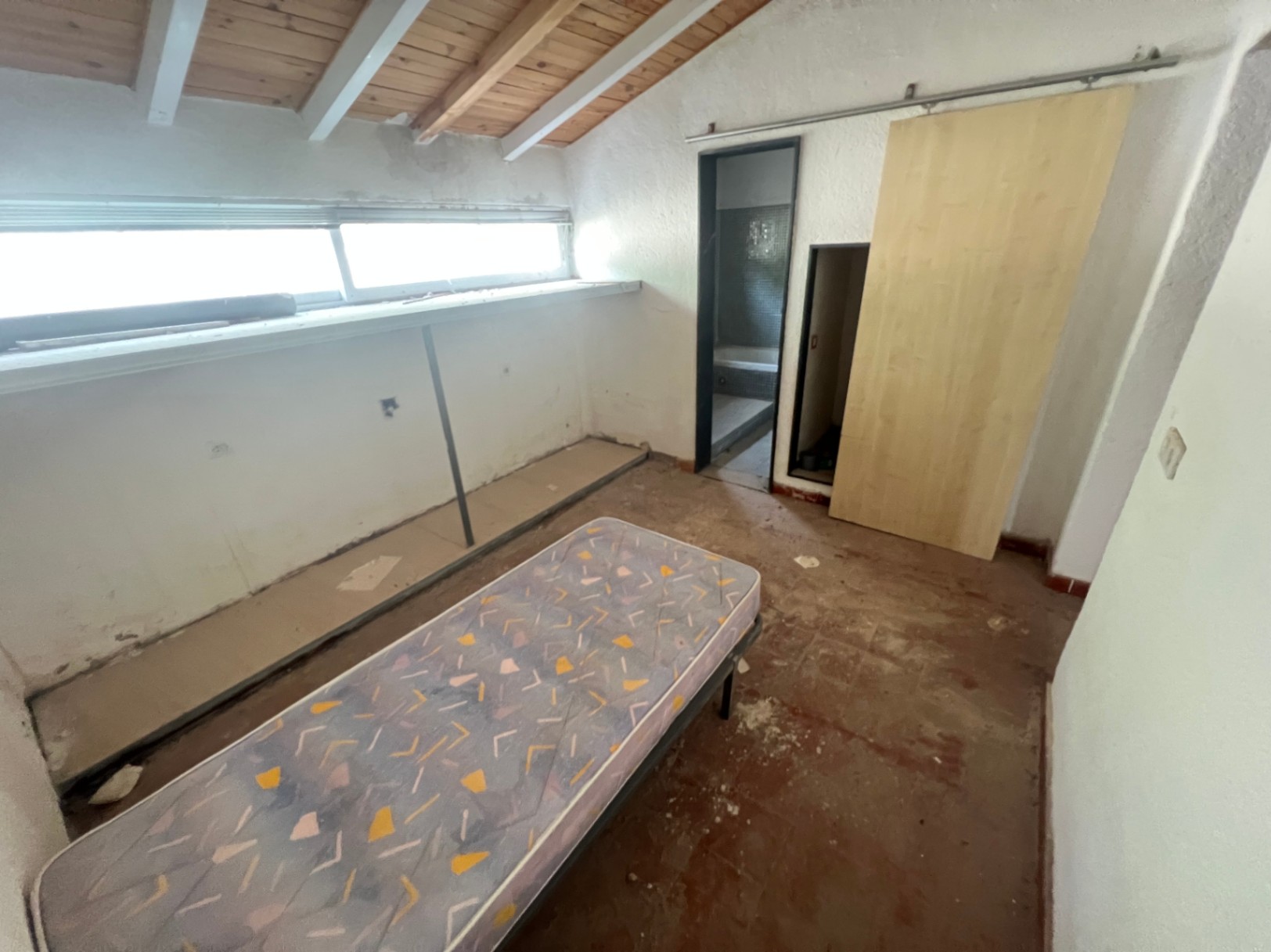 Dormitorio con techos de vigas de madera vista, ventana longitudinal y acceso a baño privado. Espacio diáfano que requiere reforma integral.