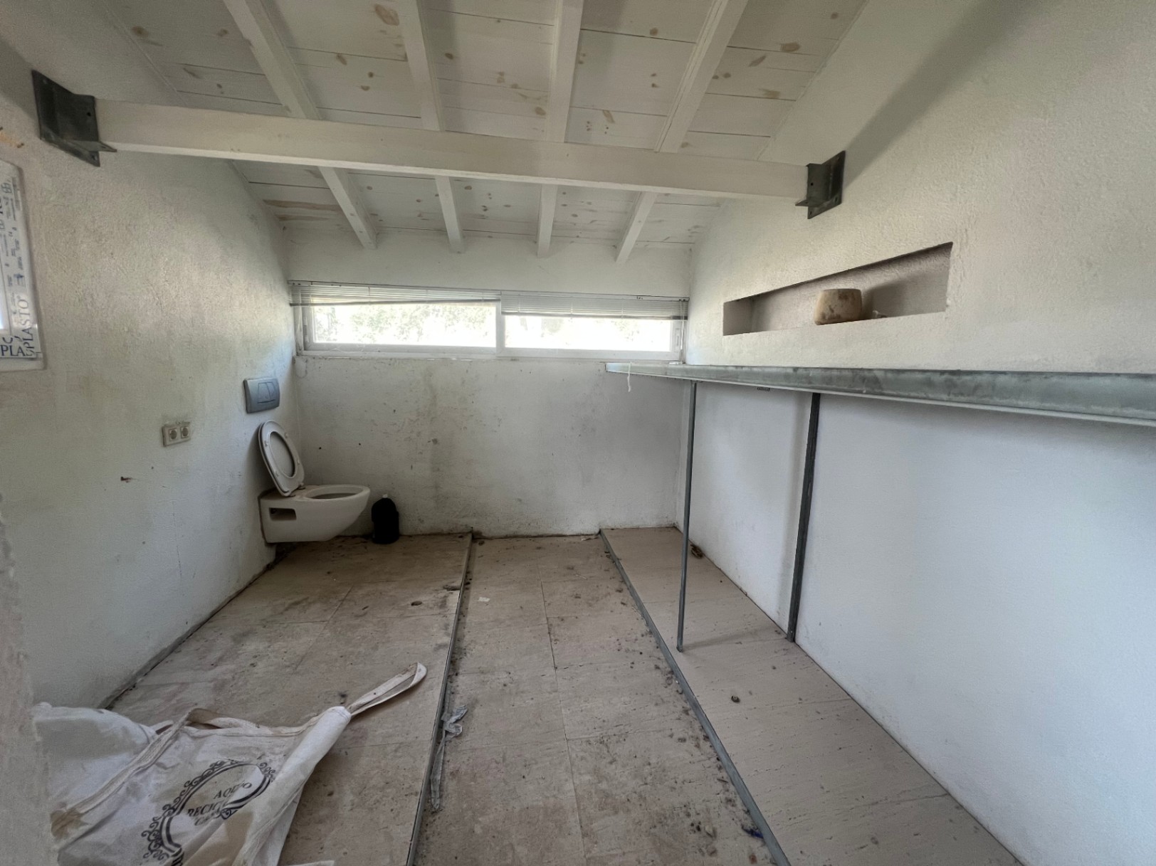 Espacio interior para reformar con techos de vigas de madera blancas, ventana horizontal, inodoro y estantería metálica. Ofrece gran potencial como estudio o trastero.