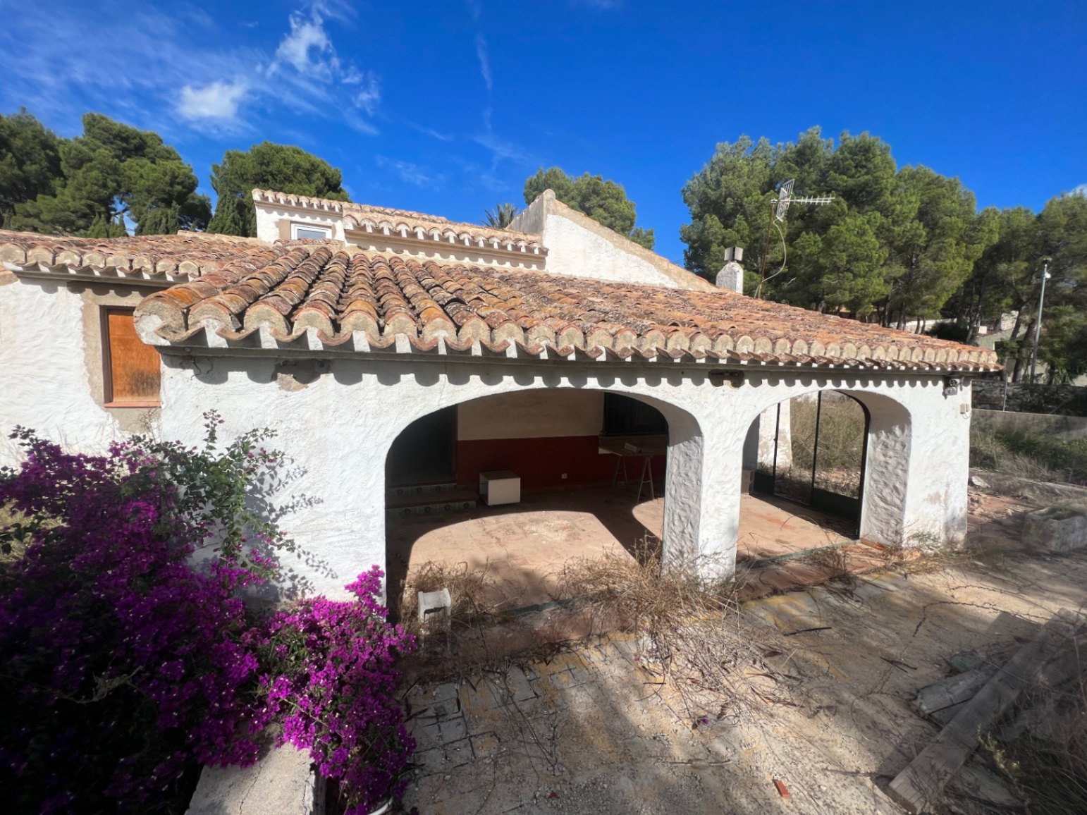 Fachada exterior con naya de arcos tradicionales, tejado de teja cerámica y paredes blancas. Destaca la buganvilla en flor y el entorno arbolado bajo un cielo despejado.
