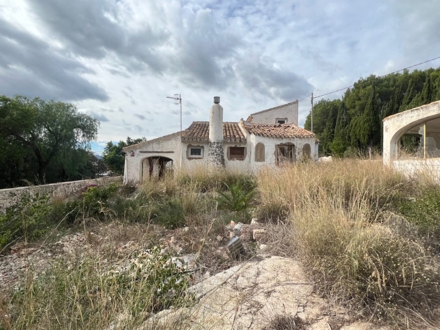 Fachada exterior de estilo tradicional con chimenea de piedra y tejado de teja, rodeada de una amplia parcela con vegetación autóctona y pinos en Jávea.