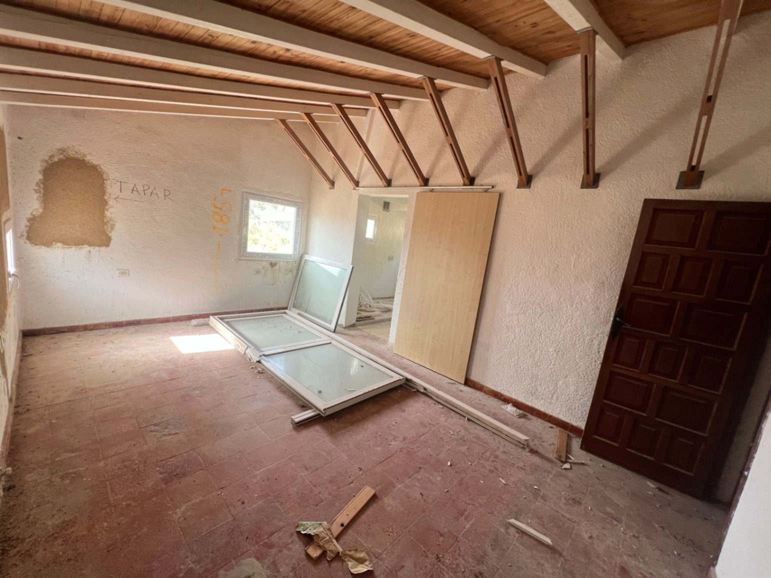 Habitación en proceso de reforma con techos de vigas de madera vistas, suelos de terracota originales y carpintería exterior pendiente de instalación.
