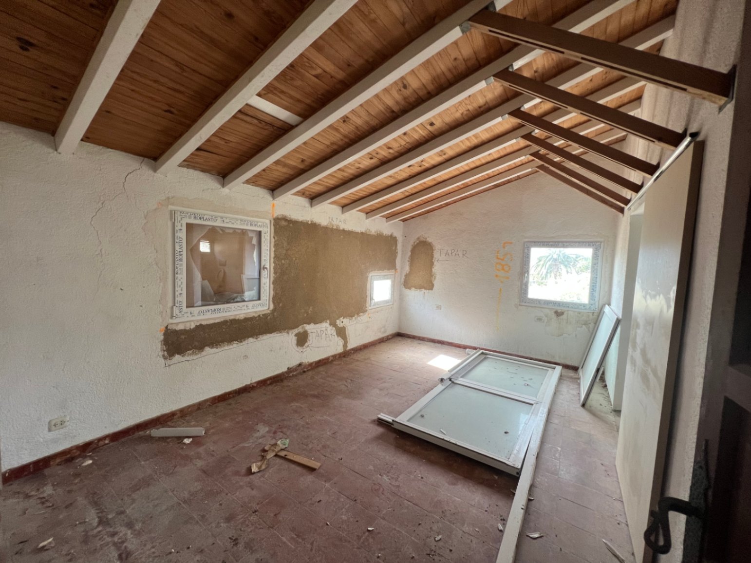 Habitación en proceso de reforma con techos de vigas de madera vistas, suelos de terracota y nuevas carpinterías de PVC ya integradas.