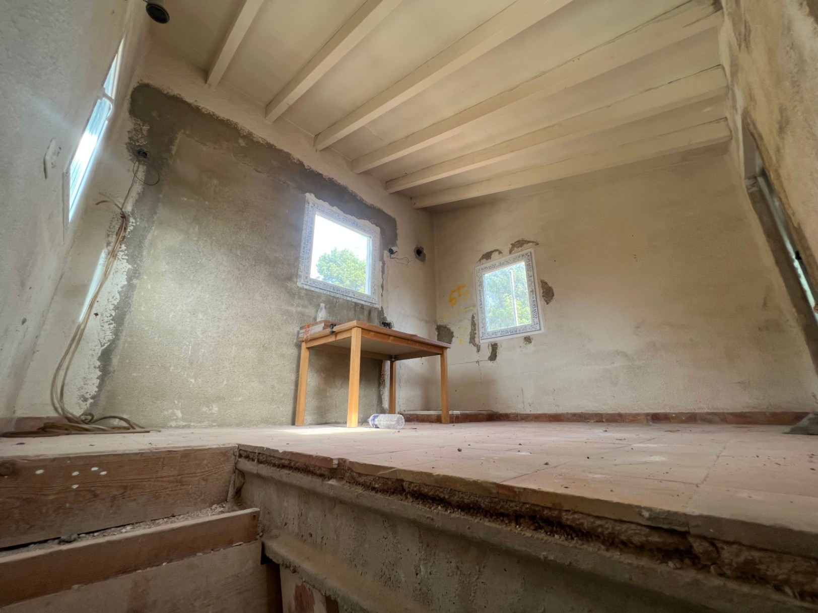 Habitación en proceso de reforma integral con vigas de madera vistas, suelos de terracota y paredes de hormigón. Dispone de ventanas nuevas que aportan luz natural.