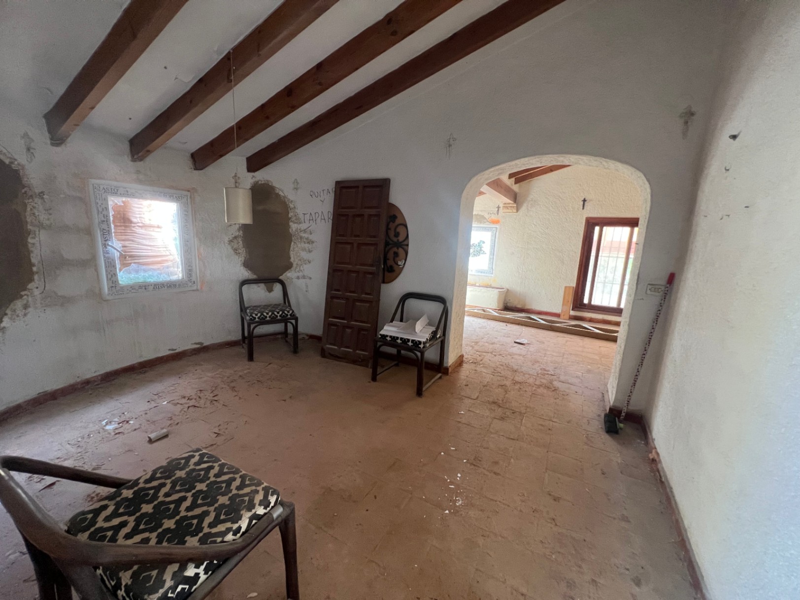 Interior con techos de vigas de madera vistas, suelos de barro y un arco que comunica las estancias. Espacio diáfano en proceso de reforma con luz natural y elementos rústicos.