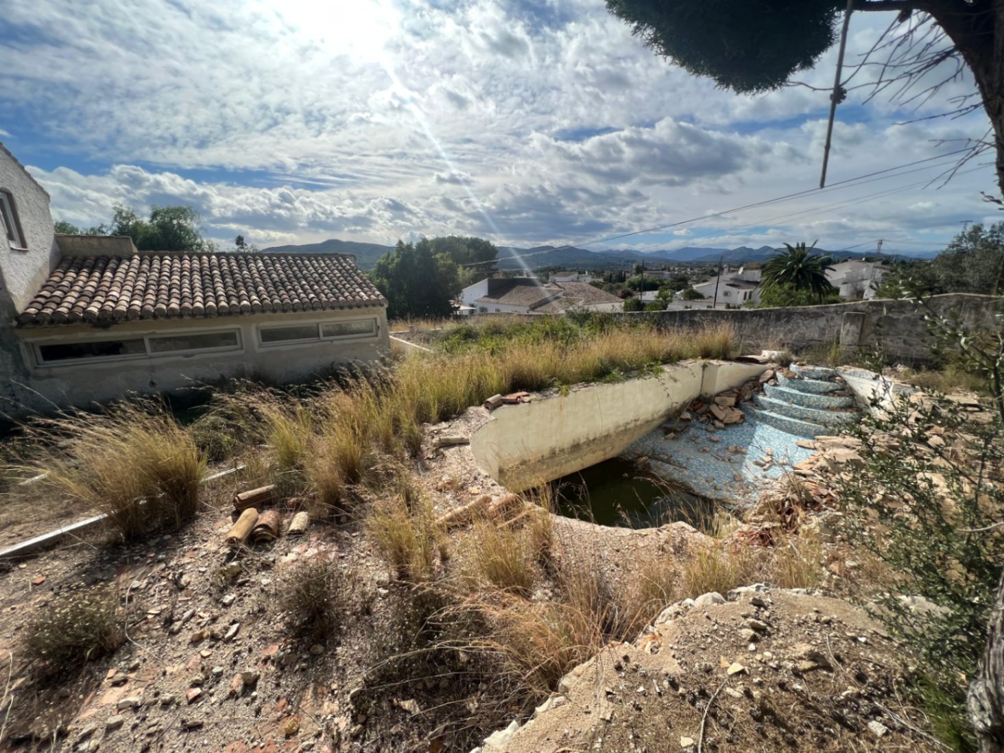 Piscina para reformar con escaleras romanas, rodeada de vegetación natural y vistas abiertas a las montañas del valle de Jávea bajo un cielo despejado.