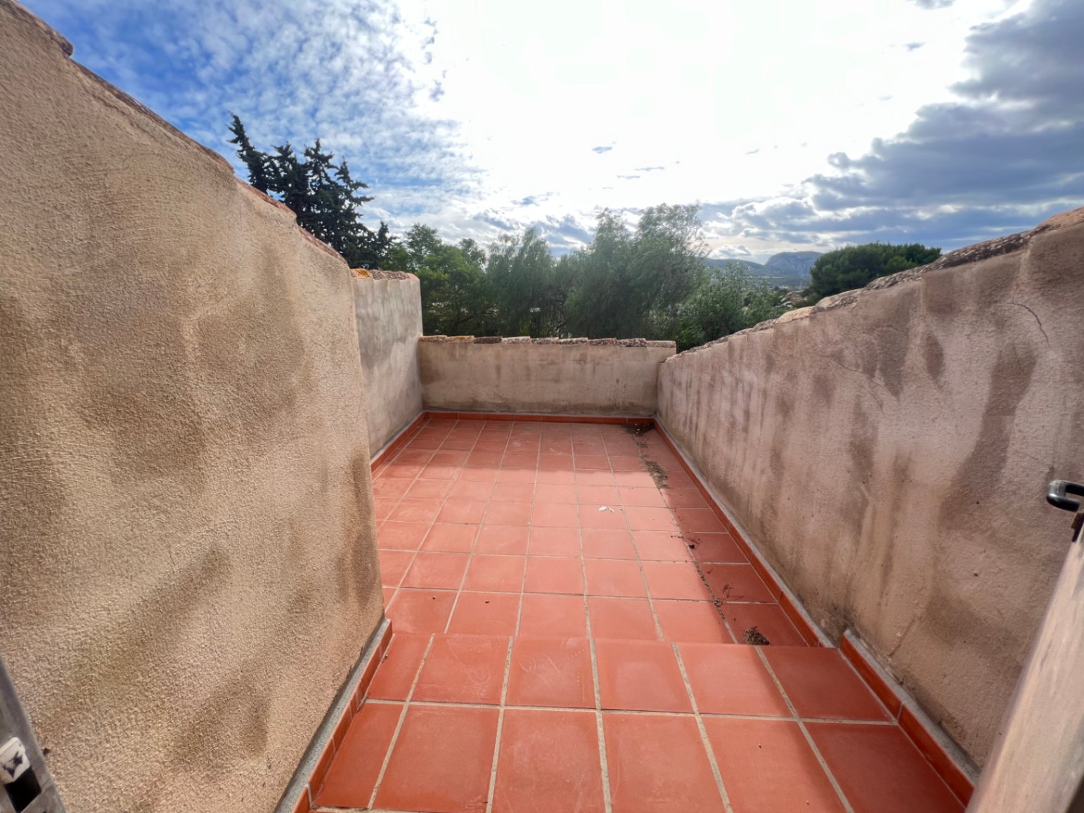 Terraza superior con suelo de baldosas de barro cocido y muros altos enfoscados, ofreciendo vistas despejadas a la vegetación circundante y las montañas de Jávea.