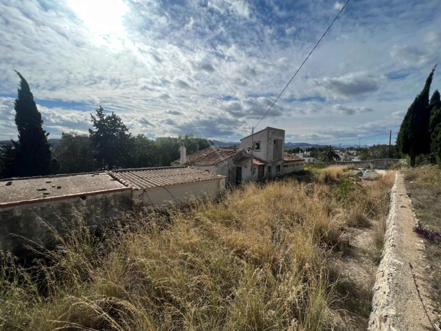 Vista exterior de la construcción original y anexos sobre la amplia parcela, rodeada de vegetación natural y muros de piedra bajo un cielo despejado en Jávea.