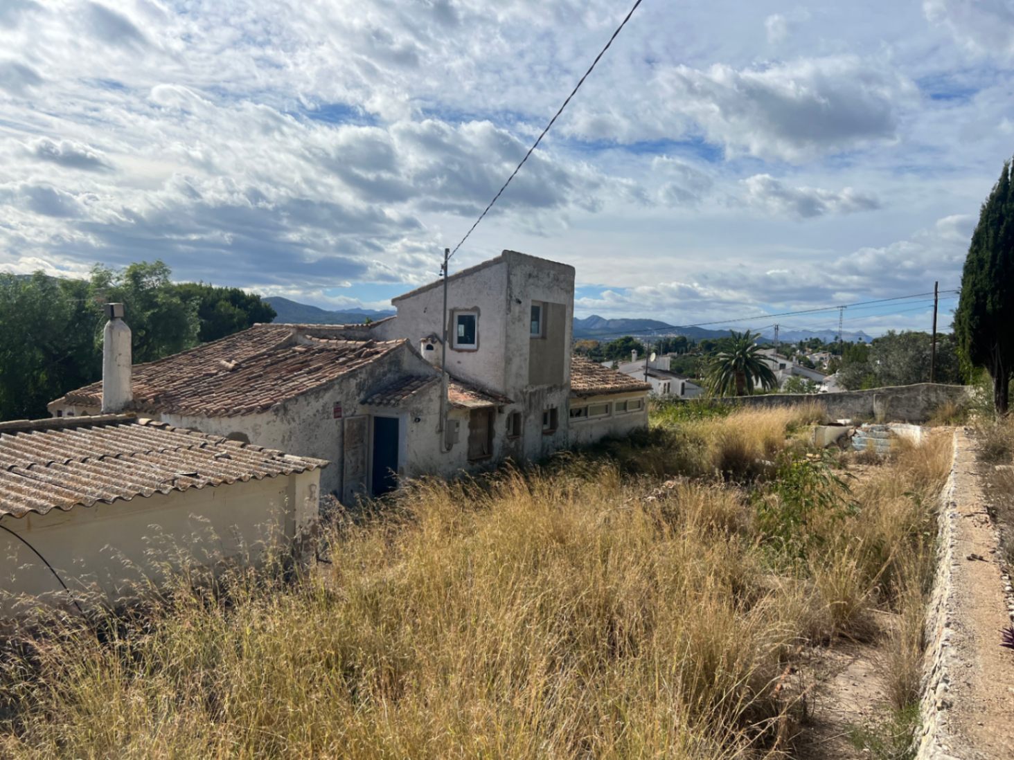 Vista exterior de la vivienda tradicional con tejados de teja y muros de estuco, rodeada de una amplia parcela con vegetación silvestre y vistas a la montaña.
