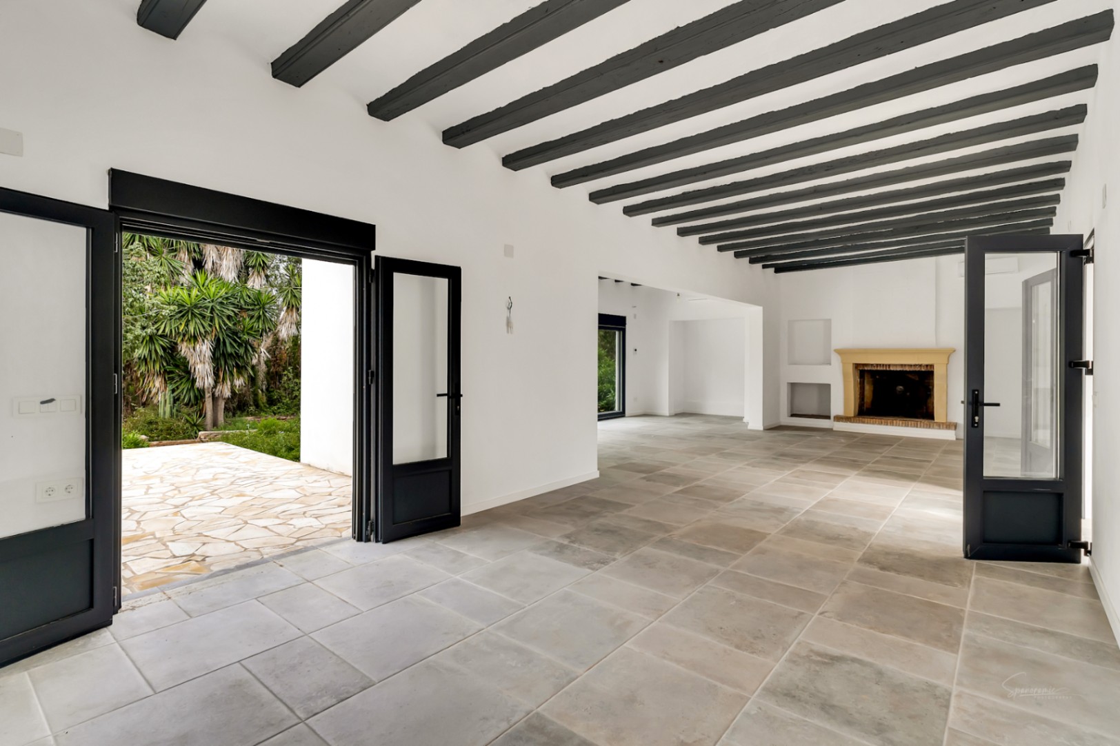 Amplio salón con vigas de madera oscura a la vista, suelos de piedra natural y chimenea clásica. Grandes ventanales de carpintería negra conectan el interior con el jardín.