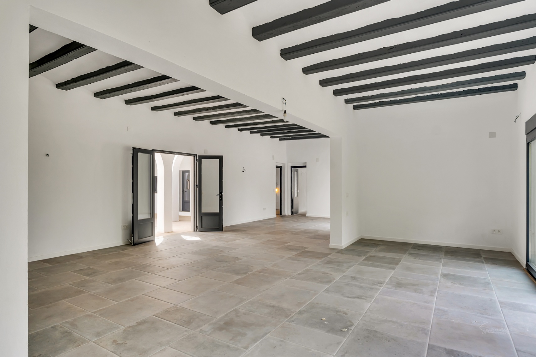 Amplio salón diáfano con suelos de piedra natural y techos altos decorados con vigas de madera oscura. Grandes puertas acristaladas conectan el espacio con el patio interior.