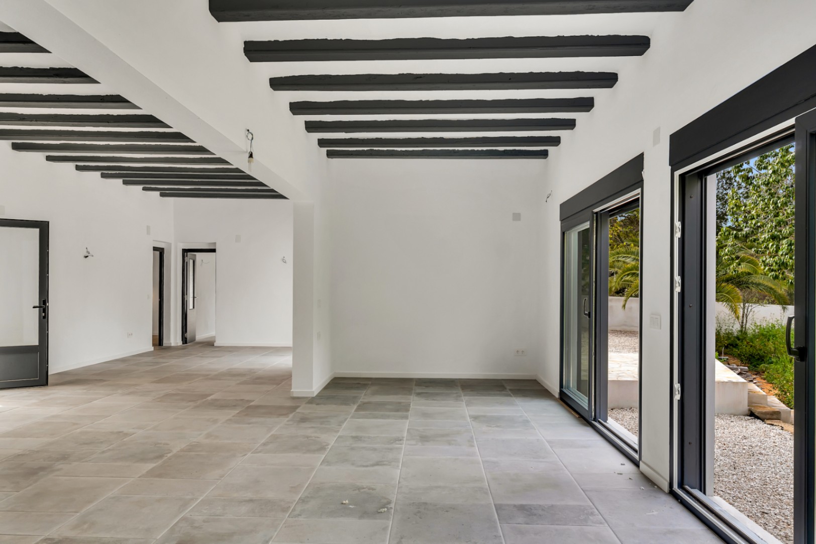 Amplio salón diáfano con vigas de madera oscura vistas, suelos de piedra de gran formato y grandes ventanales correderos que conectan con el jardín exterior.