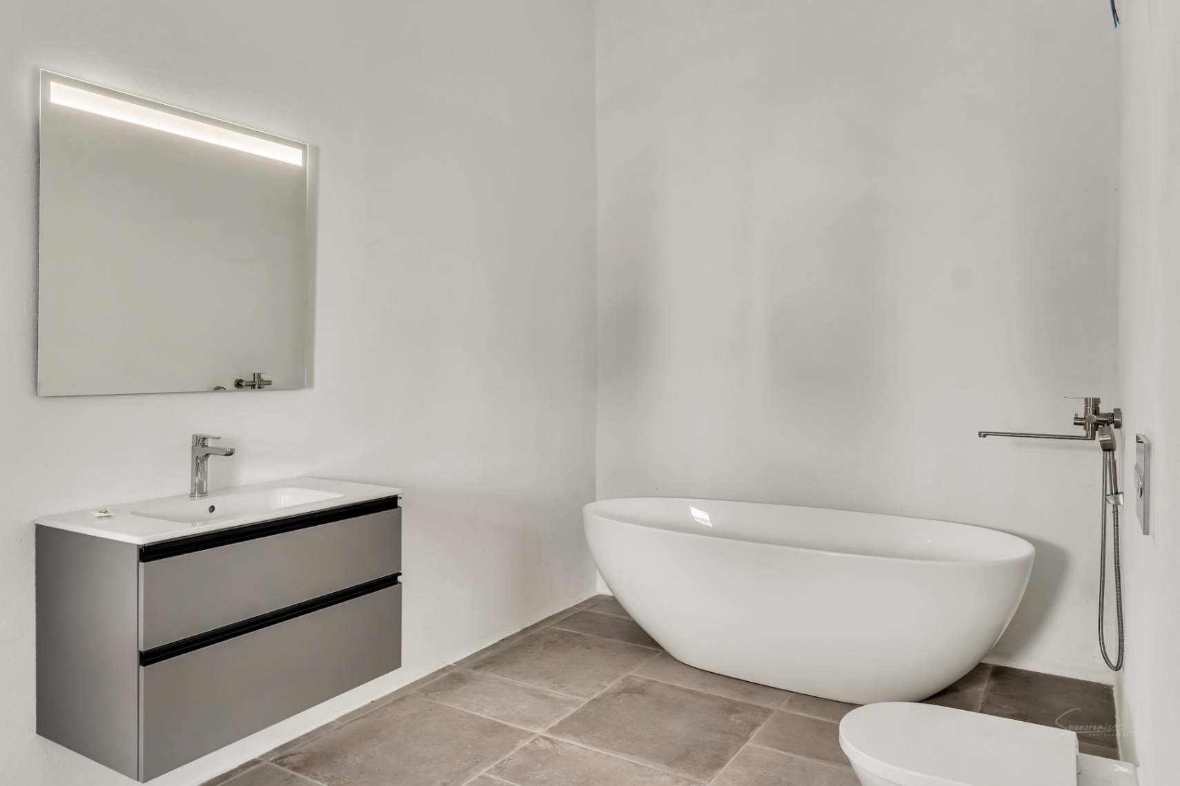Baño moderno con bañera exenta ovalada, lavabo suspendido en gris mate y suelos de piedra natural. El espejo retroiluminado complementa las paredes de estuco blanco.