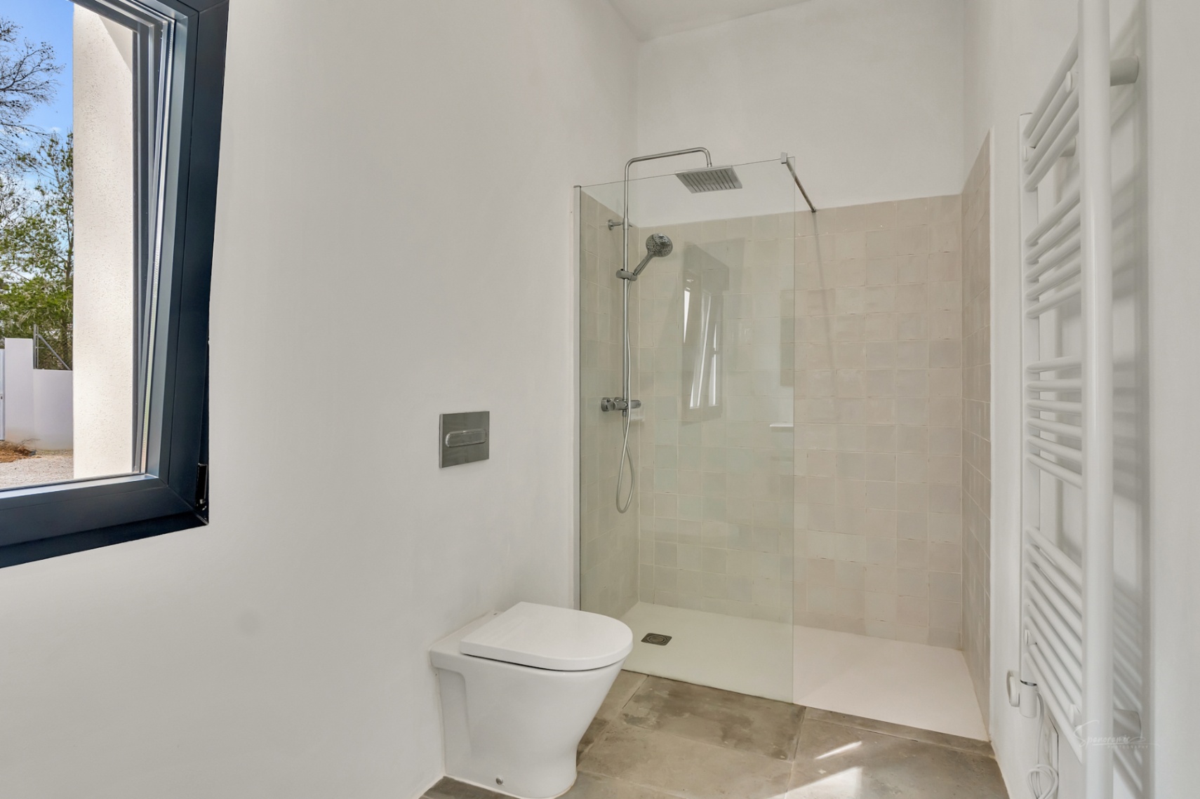 Baño moderno con ducha a ras de suelo, mampara de cristal y grifería cromada. Incluye inodoro suspendido, radiador toallero blanco y suelos de piedra natural.