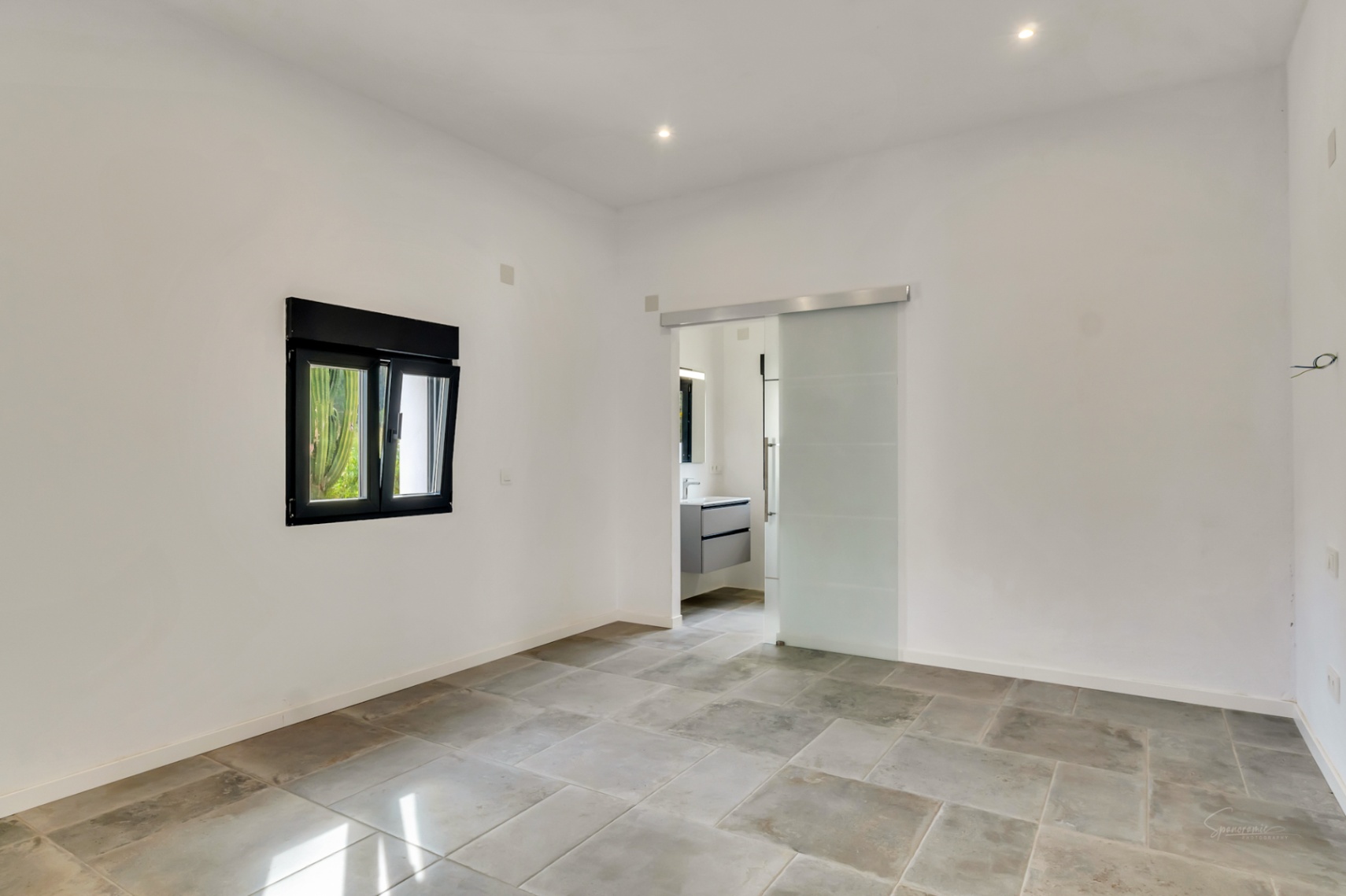 Dormitorio con suelos de piedra natural, iluminación empotrada y acceso a baño en suite mediante una puerta corredera de cristal translúcido.