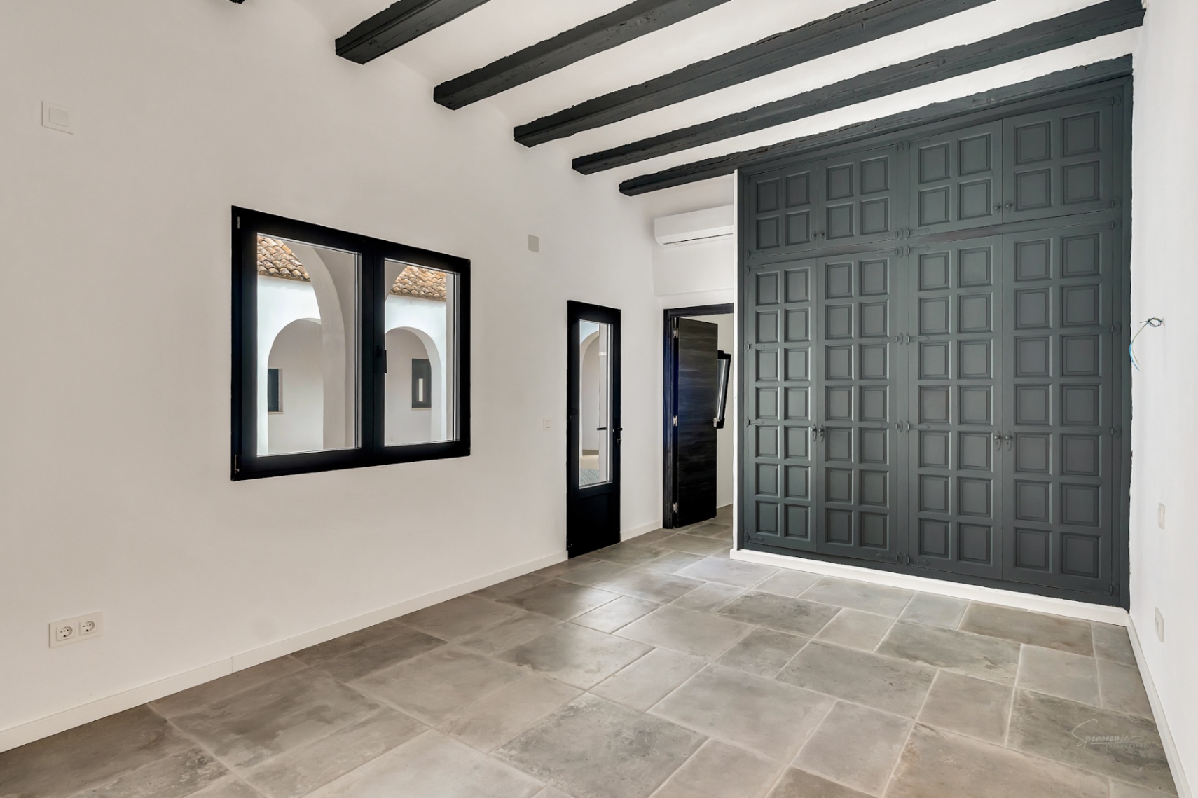 Dormitorio con vigas de madera oscuras, suelos de piedra gris y un gran armario empotrado de diseño cuadriculado. La ventana ofrece vistas al patio interior de arcos blancos.