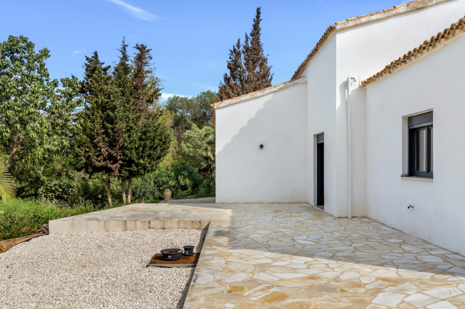 Exterior de la villa con fachada de estuco blanco, tejado de terracota y una amplia terraza de piedra natural rodeada de vegetación mediterránea.