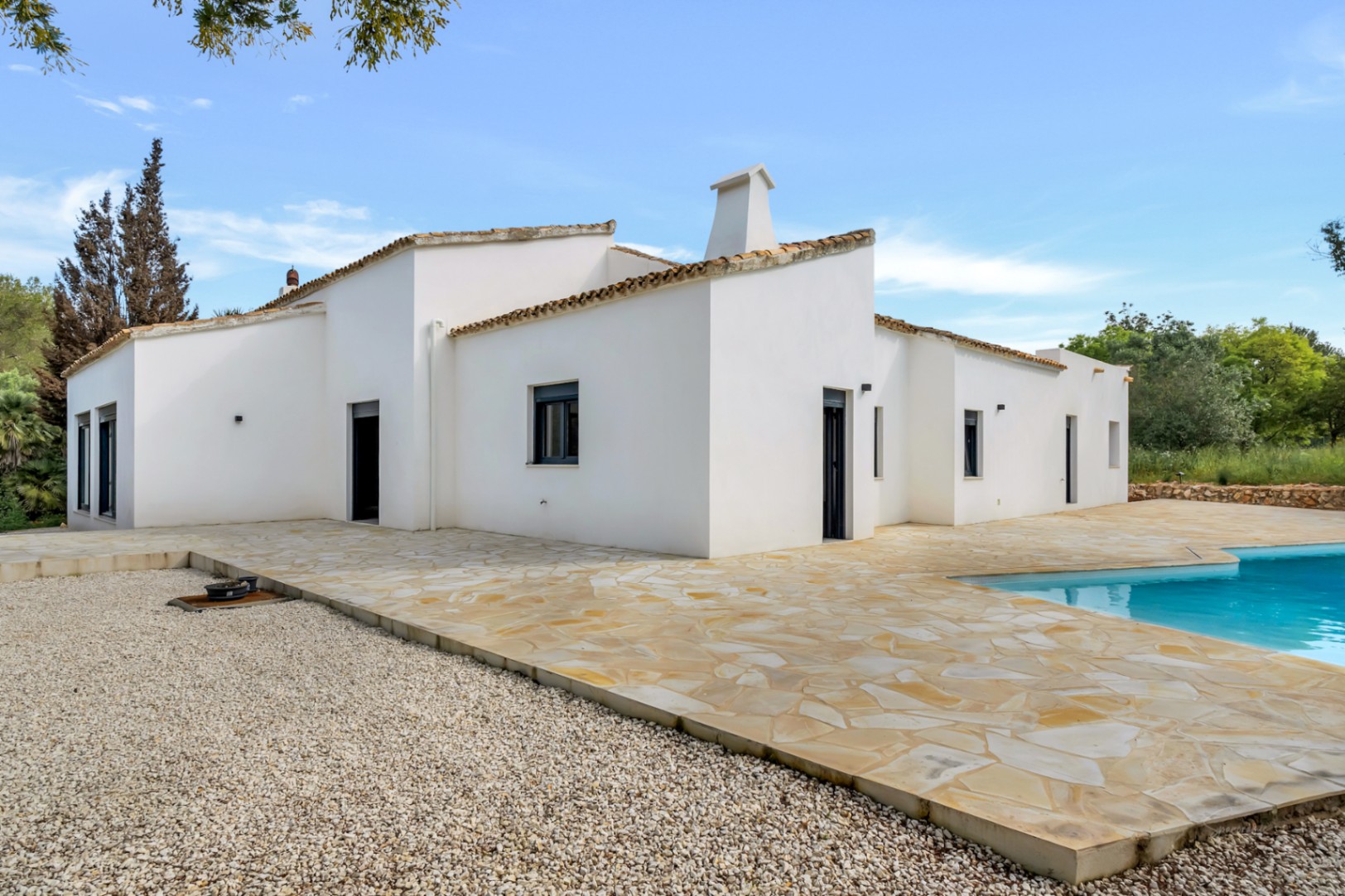 Exterior de la villa de una planta con fachada blanca, amplia terraza de piedra irregular y zona de piscina bajo el cielo despejado.