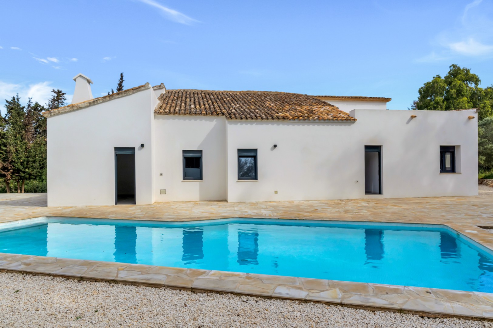 Fachada blanca de estilo mediterráneo con tejado de teja y carpintería oscura frente a una piscina rodeada de terraza de piedra natural y zona de grava.