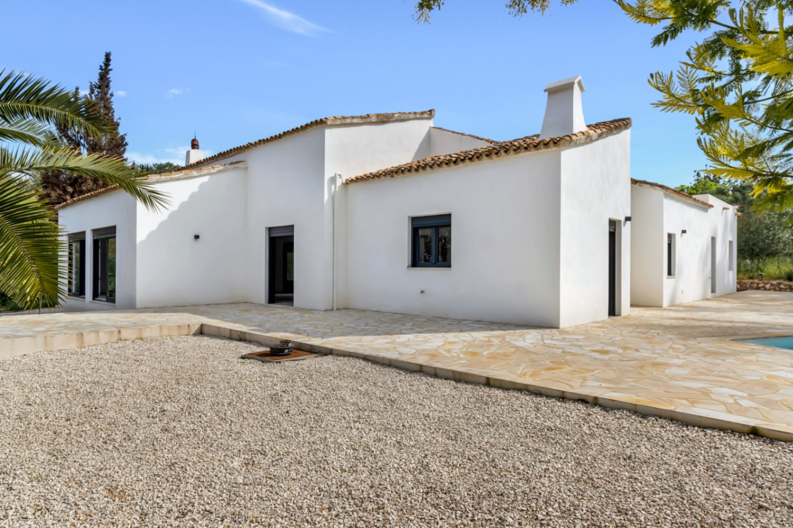Fachada exterior de estilo mediterráneo con paredes de estuco blanco, techos de teja y una amplia terraza de piedra natural que conecta con una zona de grava.