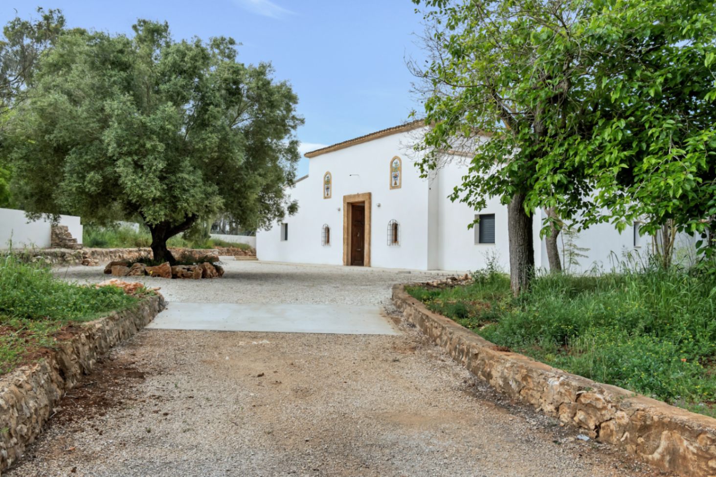 Fachada principal de estilo mediterráneo con muros de estuco blanco, entrada de piedra natural y un amplio camino de acceso rodeado de vegetación y olivos centenarios.
