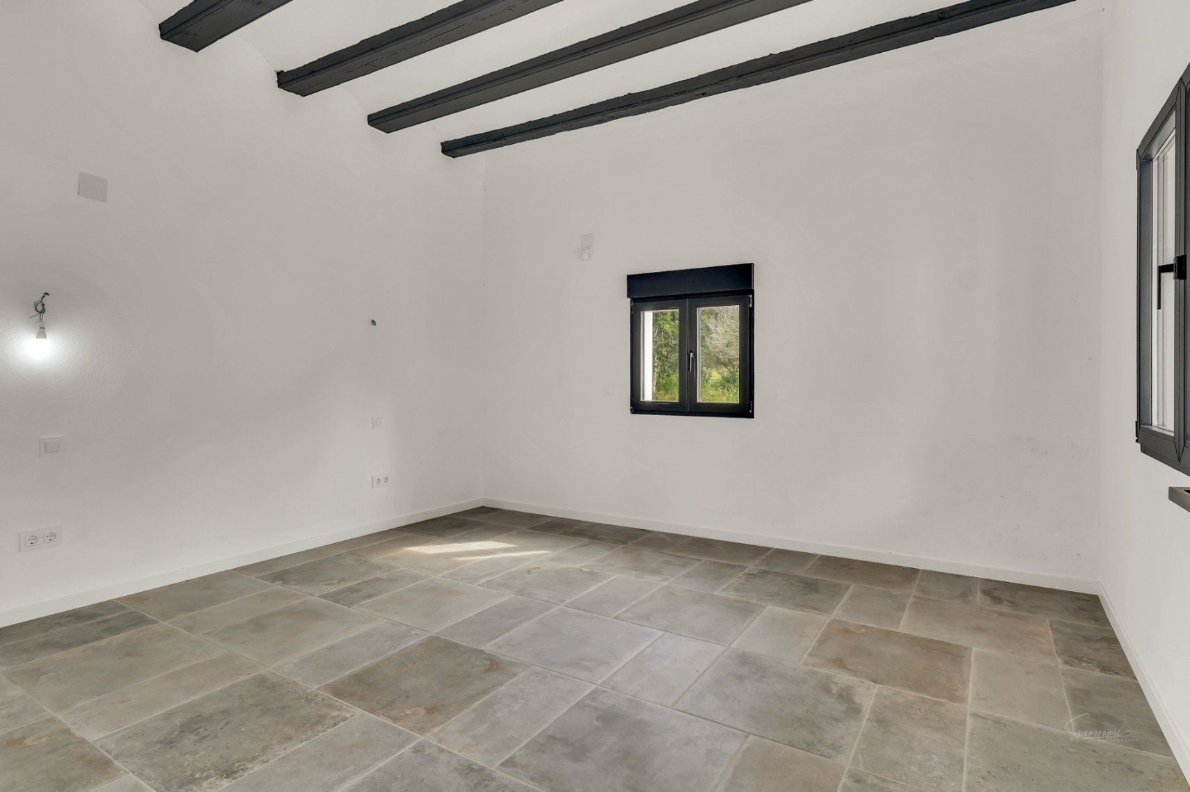 Habitación diáfana con suelos de piedra de gran formato, techos altos con vigas de madera oscura expuestas y carpintería exterior en color negro.