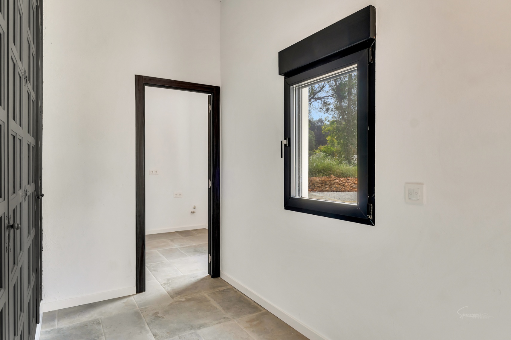Pasillo con suelos de piedra natural y paredes blancas, destacando la carpintería oscura en puertas y ventana con vistas al jardín exterior.