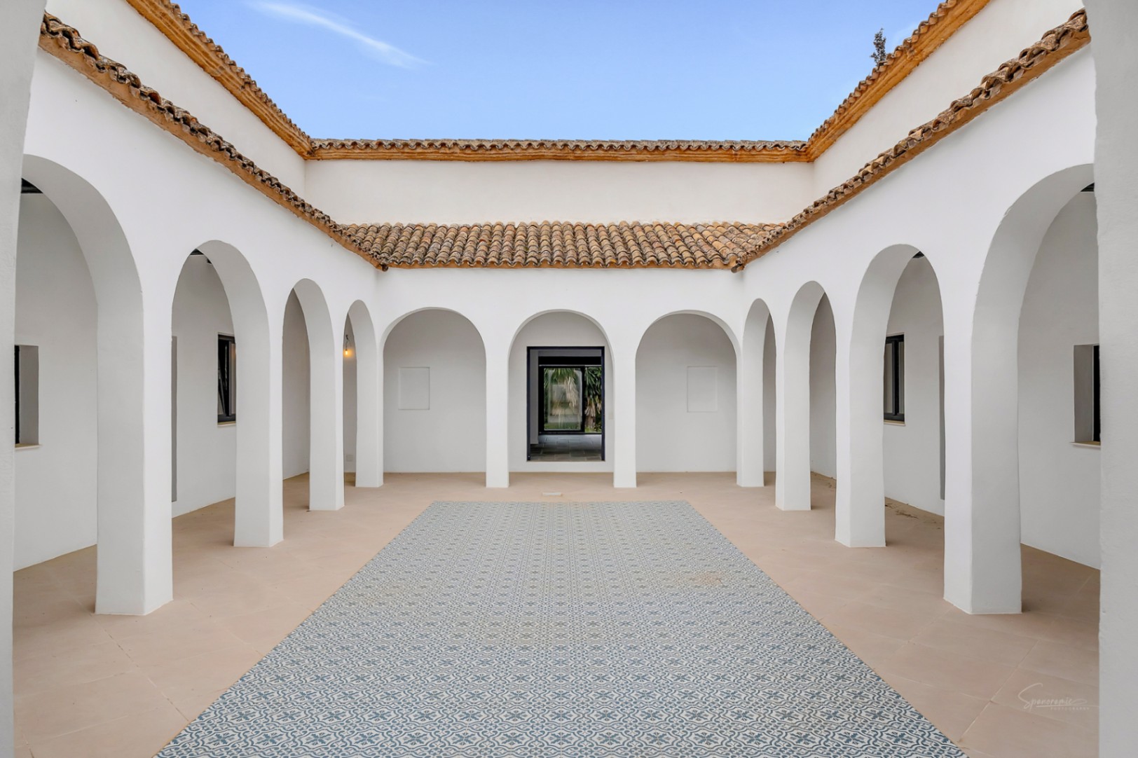 Patio interior de estilo mediterráneo con arcos blancos simétricos, suelos de piedra y una alfombra central de baldosas hidráulicas bajo un techo de teja tradicional.