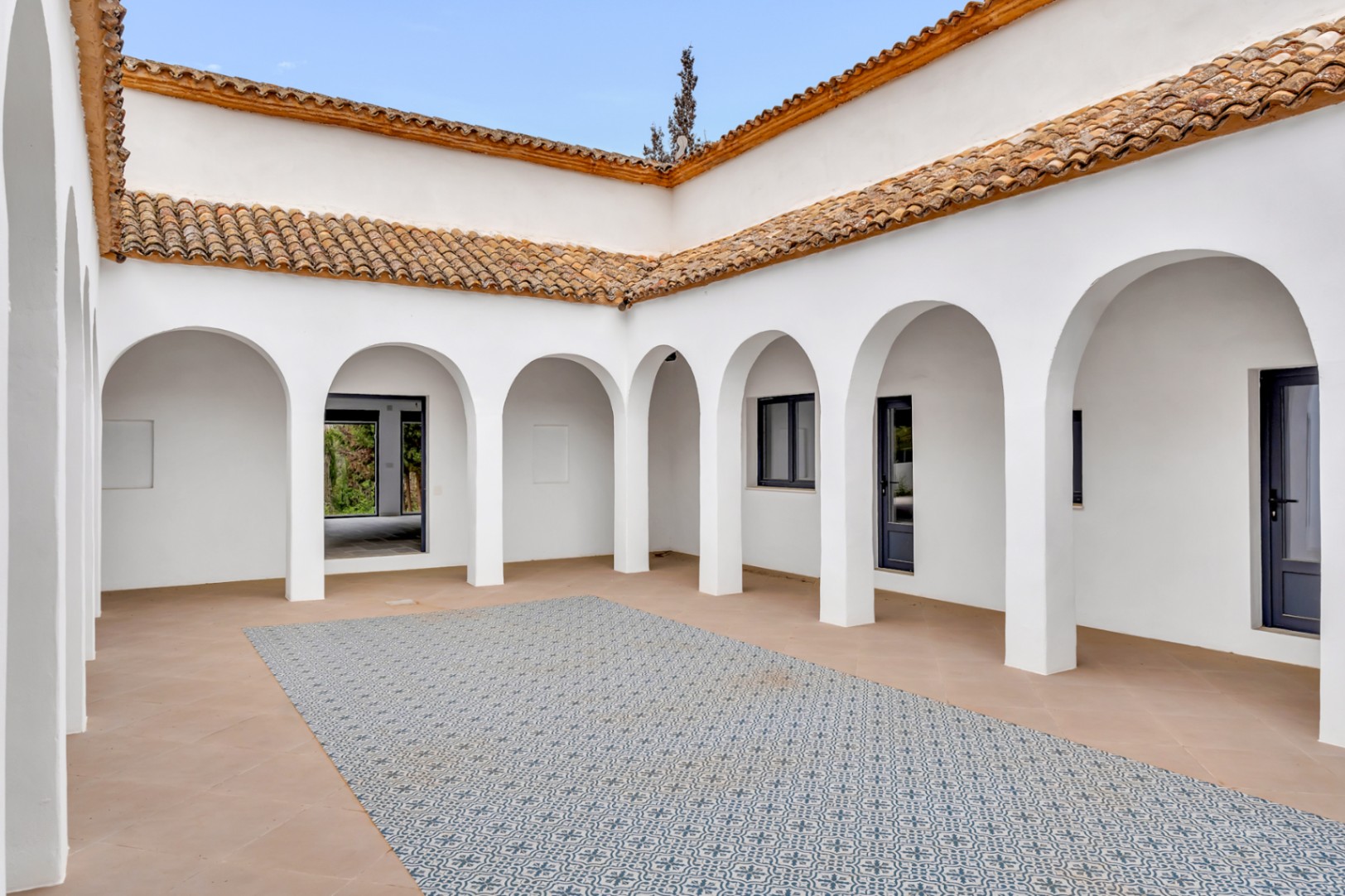 Patio interior de estilo mediterráneo con arcos blancos, suelos de terracota y un mosaico central de baldosas estampadas bajo un techo de tejas tradicionales.