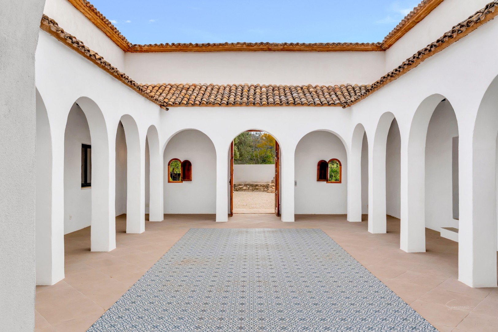 Patio interior de estilo mediterráneo con arcos de medio punto, paredes de estuco blanco y suelos de baldosas hidráulicas decorativas bajo un cielo abierto.
