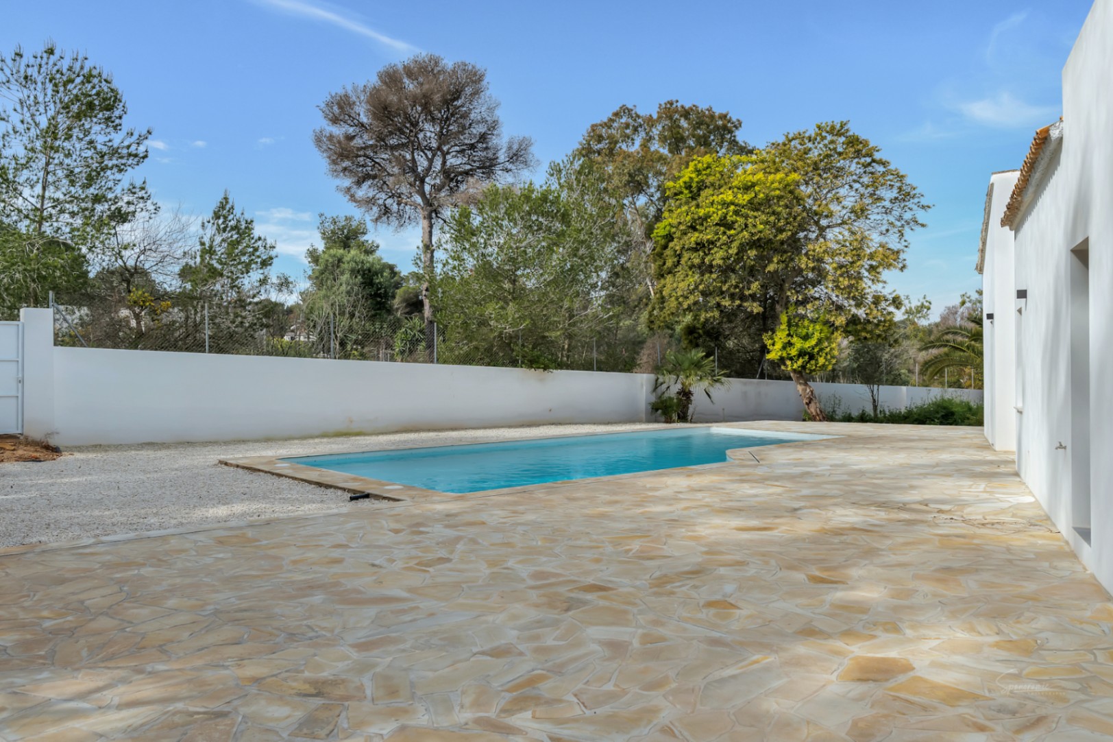 Piscina exterior rodeada por una amplia terraza de piedra natural y zona de grava, delimitada por un muro blanco y vegetación mediterránea madura.