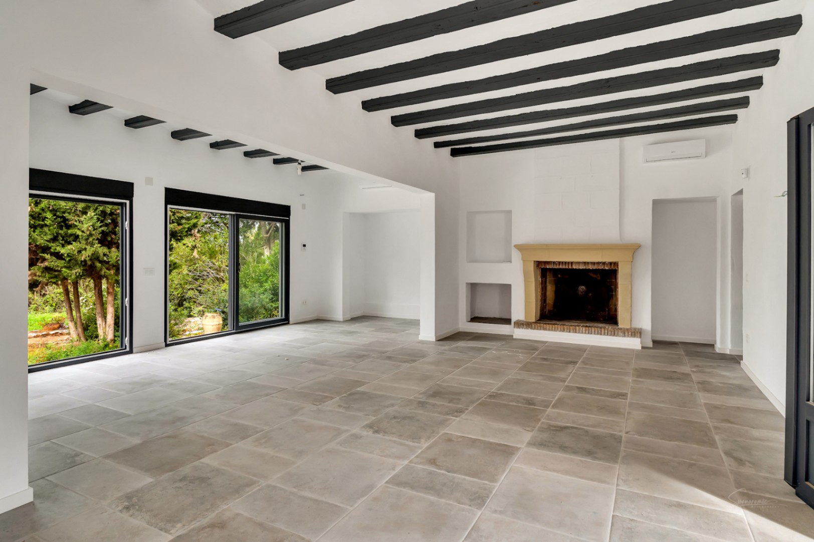 Salón diáfano con chimenea tradicional, vigas de madera oscura y suelos de piedra. Grandes ventanales correderos integran el espacio interior con las vistas al jardín.