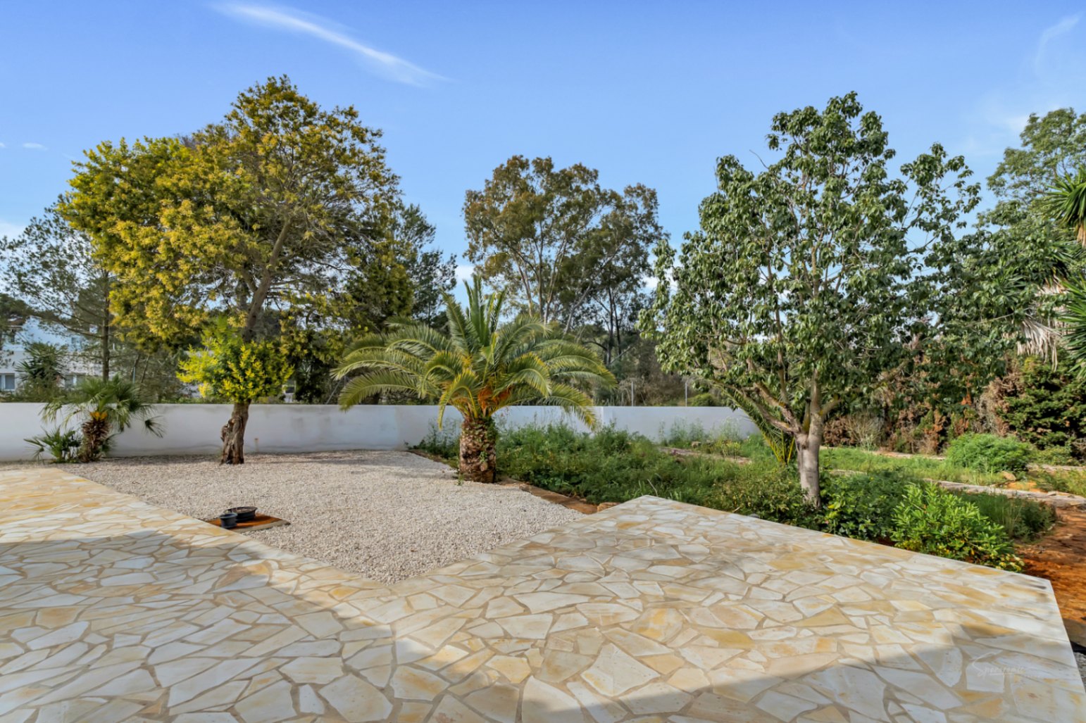 Terraza exterior con pavimento de piedra natural irregular que da paso a un jardín mediterráneo con palmeras, zonas de grava y vegetación madura bajo un cielo despejado.