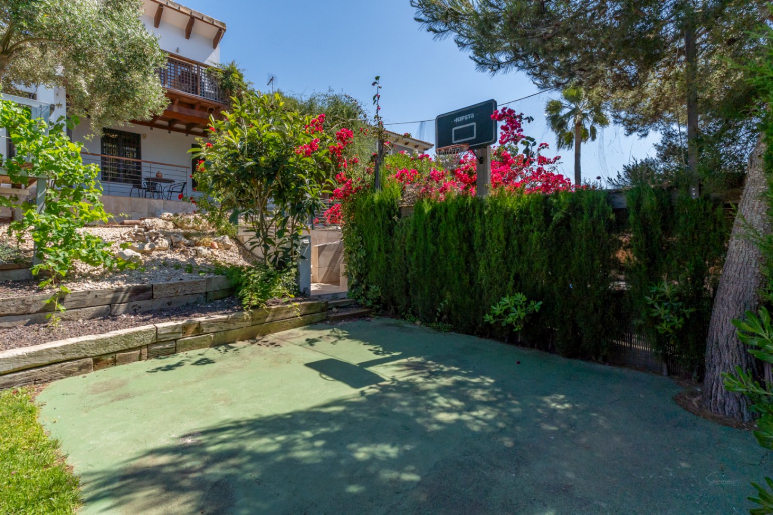 Área recreativa exterior con canasta de baloncesto, superficie verde y setos altos que garantizan privacidad, integrada en un jardín mediterráneo con buganvillas.