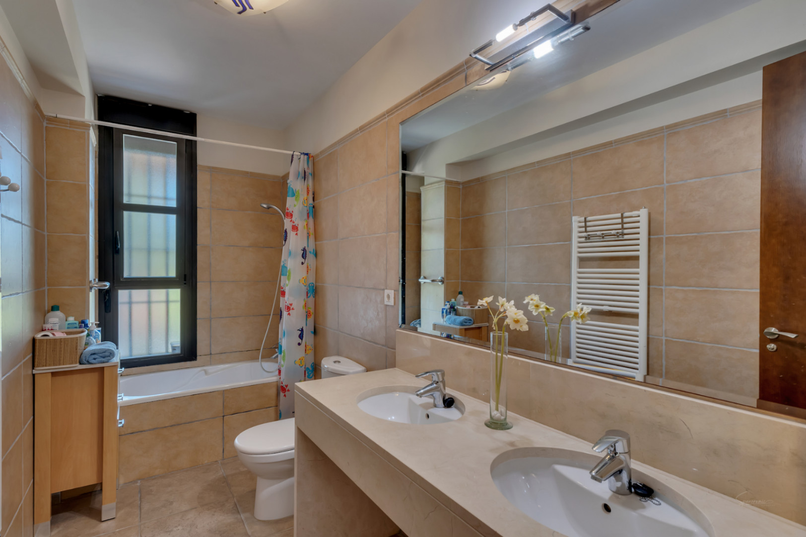 Baño completo con lavabo de doble seno, encimera de piedra y gran espejo. Cuenta con bañera, radiador toallero vertical y revestimiento cerámico en tonos beige.