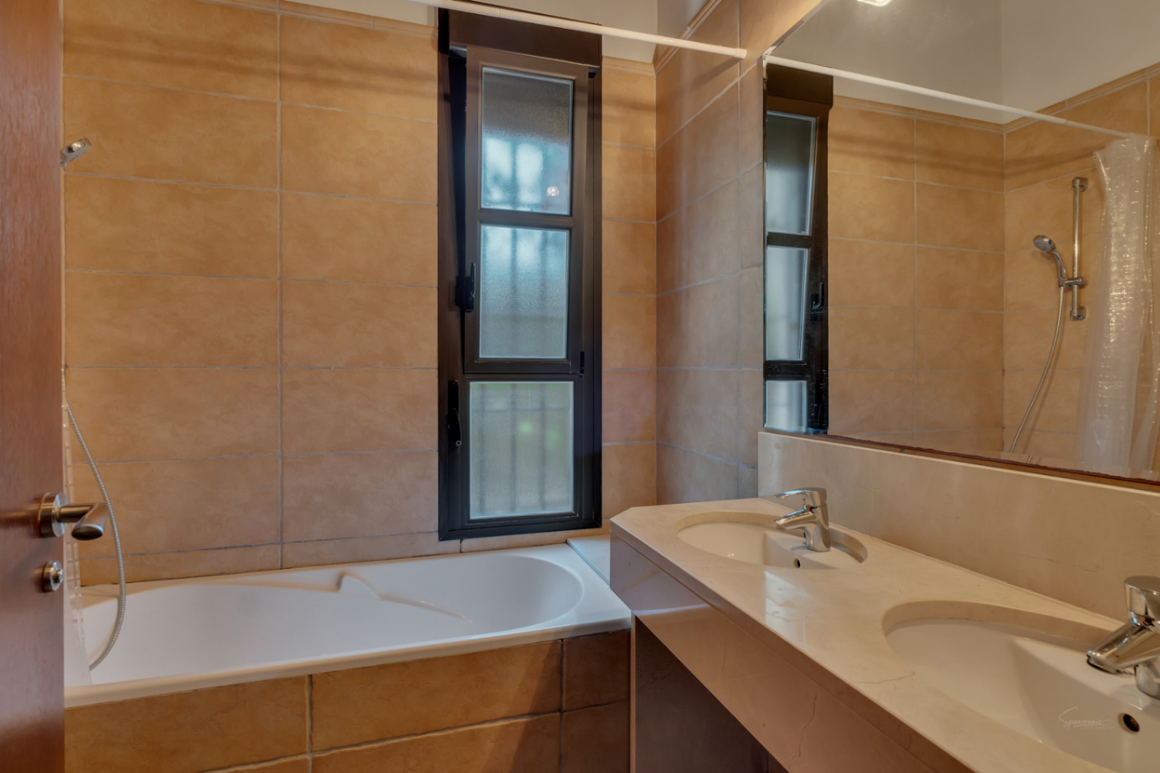 Baño con encimera de piedra de doble seno, bañera integrada y paredes revestidas en azulejos de tonos tierra junto a una ventana de marco oscuro.