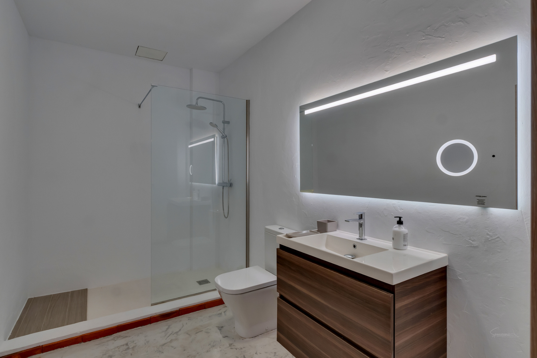 Baño moderno con mueble suspendido de madera, lavabo integrado y espejo retroiluminado. Incluye ducha a ras de suelo con mampara de cristal y paredes blancas con textura.