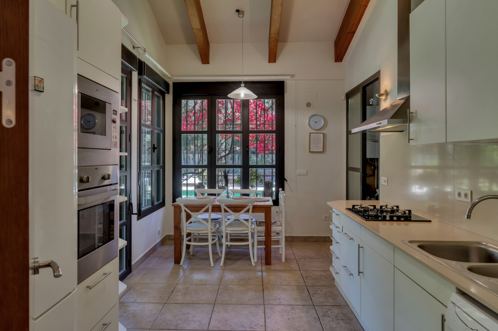 Cocina funcional con techos altos y vigas de madera, zona de comedor integrada y grandes ventanales que ofrecen vistas directas al jardín mediterráneo.