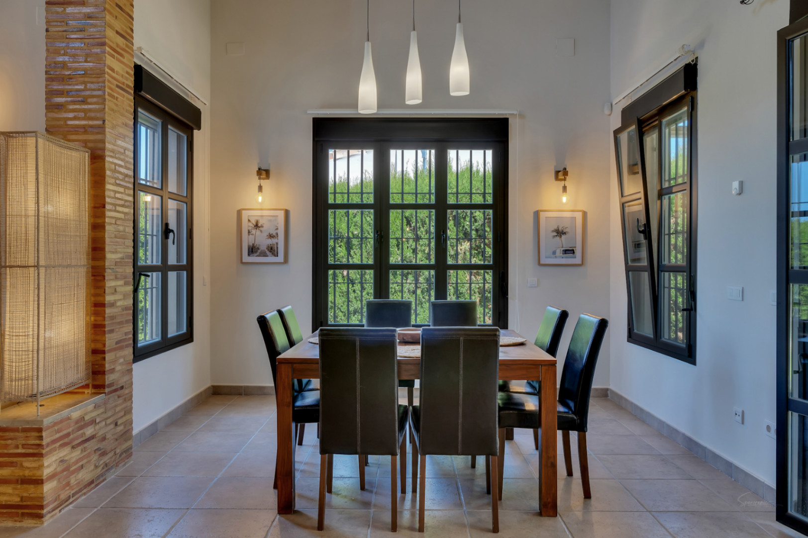 Comedor con mesa de madera y sillas de cuero negro bajo techos altos. La estancia cuenta con ventanales de carpintería oscura y un pilar lateral de ladrillo visto.