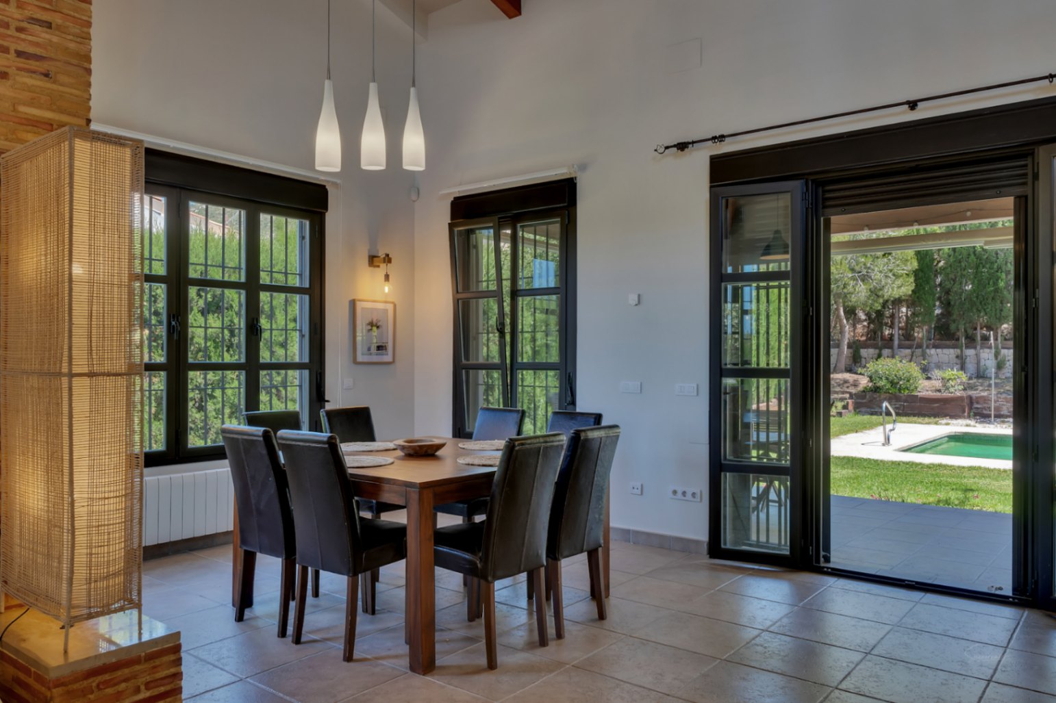 Comedor con mesa de madera y sillas de cuero oscuro, iluminado por lámparas colgantes y luz natural con vistas directas al jardín y la piscina exterior.