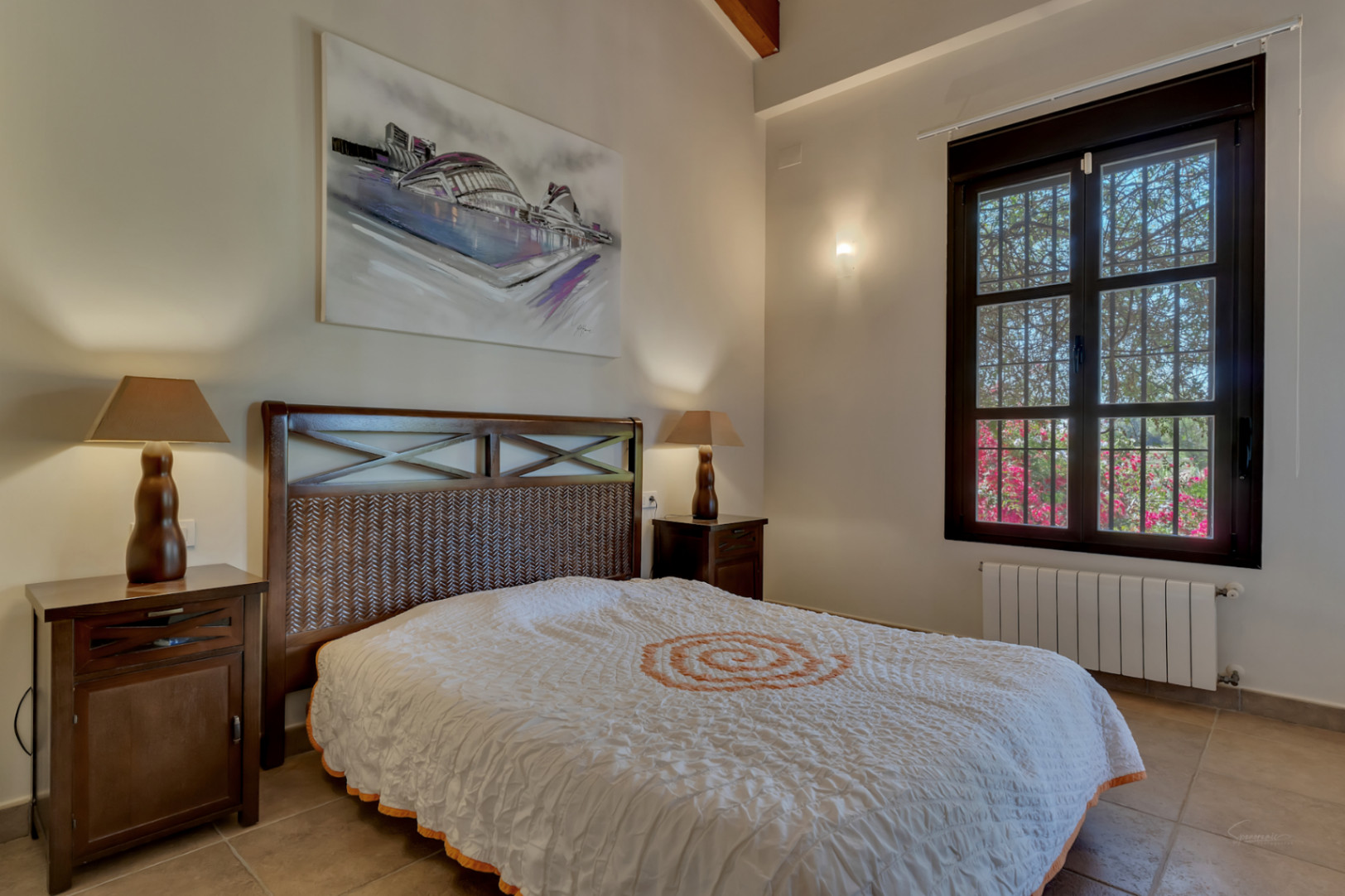 Dormitorio con cama de madera oscura y cabecero trenzado, mesitas a juego, techos altos con vigas y ventana con vistas al jardín mediterráneo.