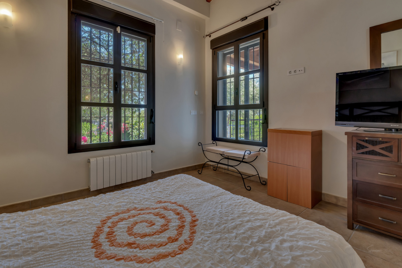 Dormitorio con ventanas de marco oscuro y rejas, vistas al jardín, calefacción por radiador, banco de forja y mobiliario de madera con televisor.