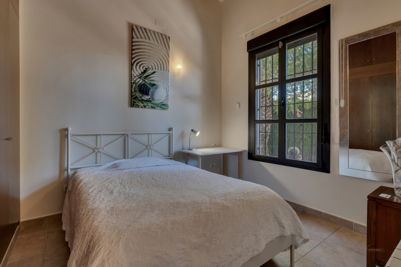 Dormitorio luminoso con cama de forja blanca, escritorio funcional y ventana de marco oscuro con vistas al jardín. Incluye suelos de gres y armario empotrado.