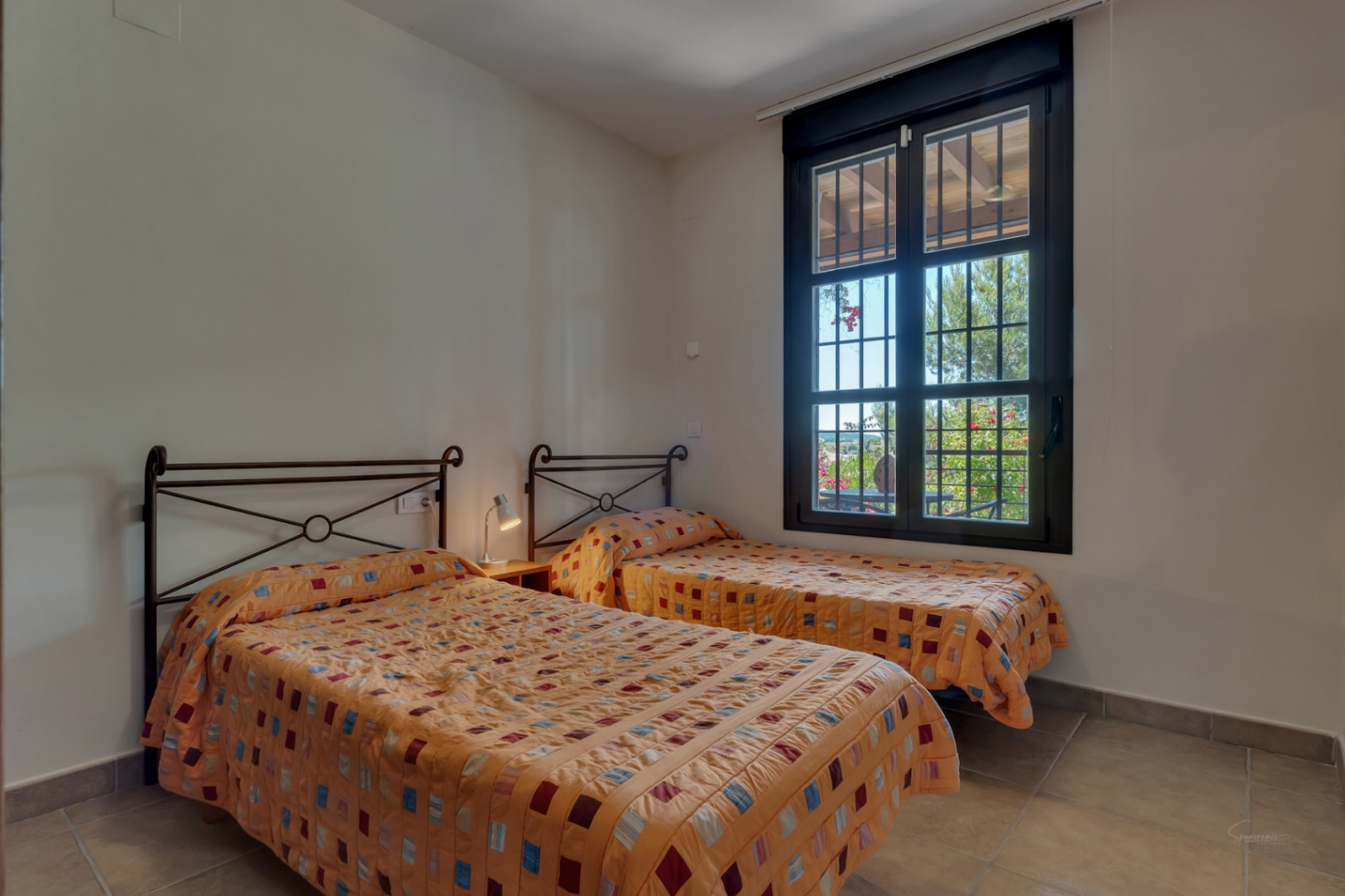 Dormitorio luminoso con dos camas individuales, cabeceros de forja y ventana con vistas al jardín y suelos de gres.