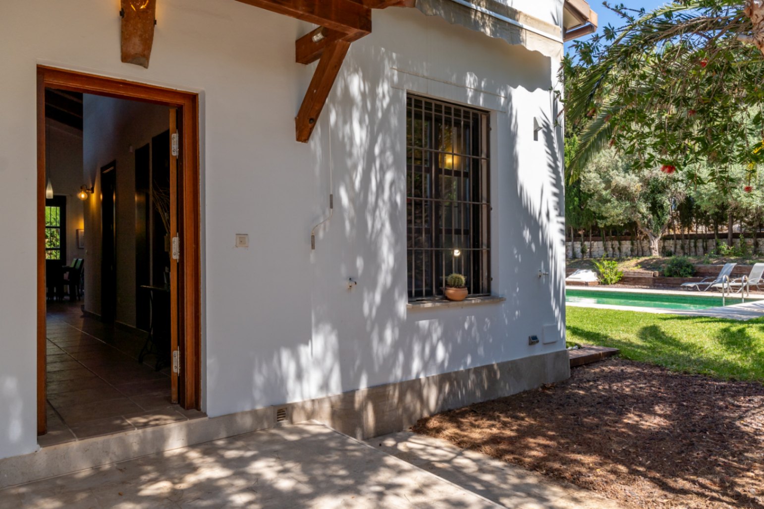 Entrada exterior con puerta de madera abierta hacia el pasillo, fachada blanca con sombras de palmeras y ventana con reja junto al jardín y la piscina.