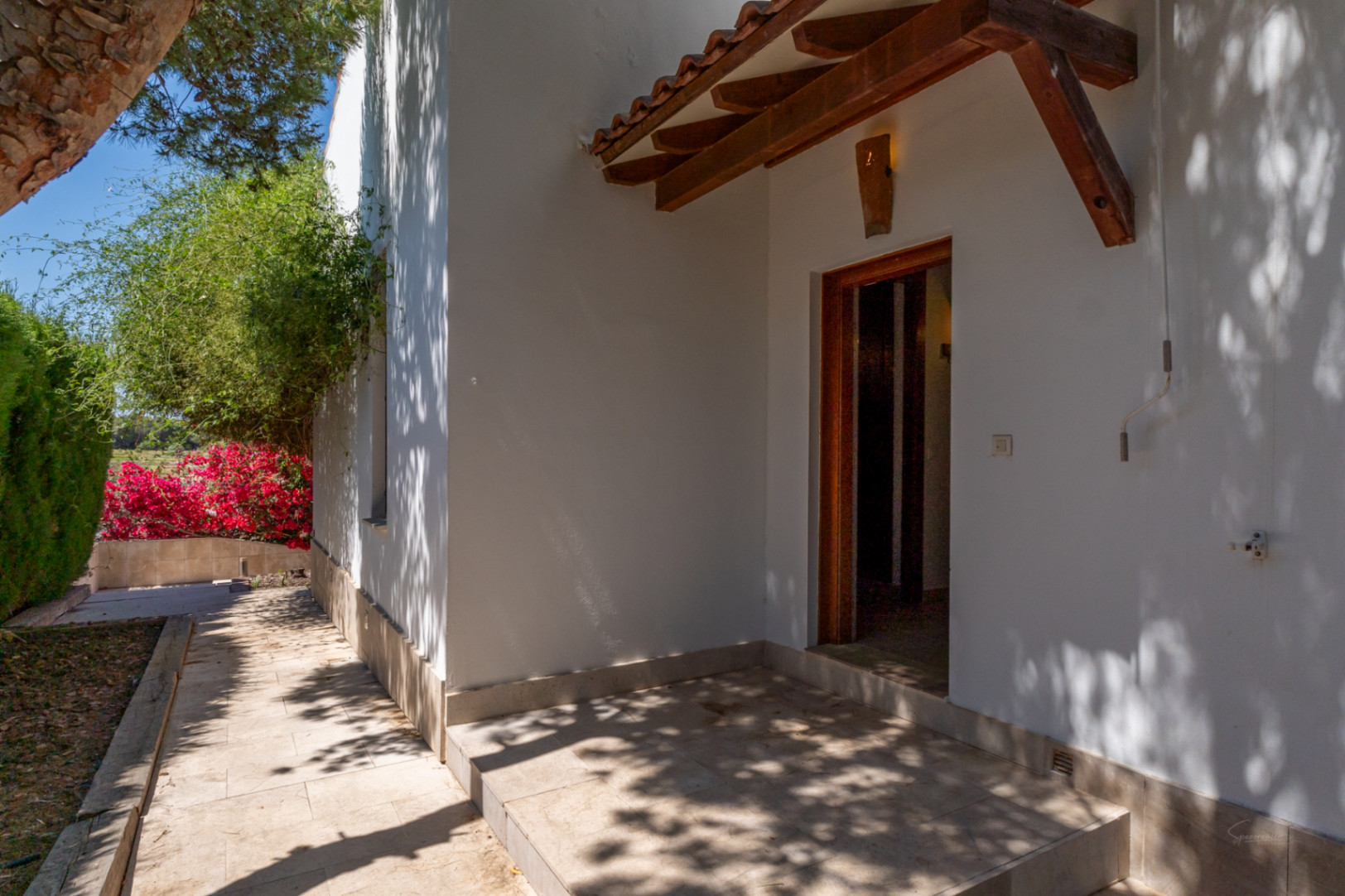 Entrada lateral con porche de madera y tejas cerámicas, destacando el suelo de piedra natural y la proximidad a la vegetación del jardín mediterráneo.