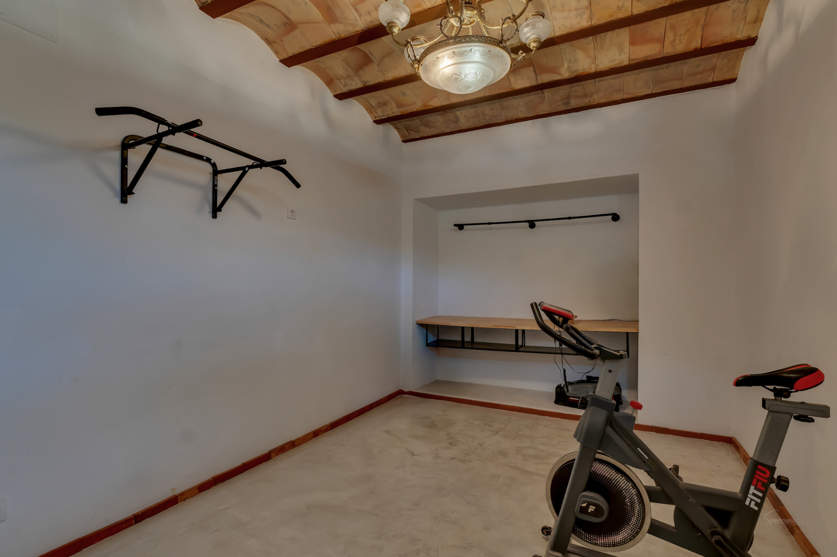Espacio polivalente con techo de bovedilla artesanal, equipado con bicicleta estática, barra de dominadas y un rincón de trabajo integrado en el nicho.