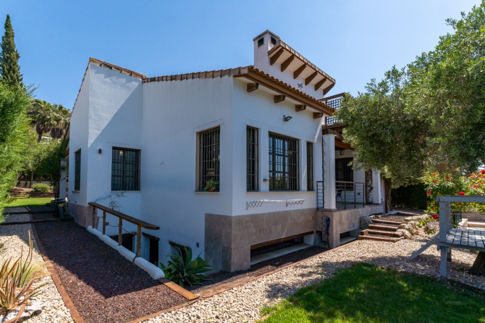 Fachada lateral de la villa con paredes blancas, vigas de madera expuestas y ventanas protegidas. Incluye acceso al semisótano y jardín con grava y césped.