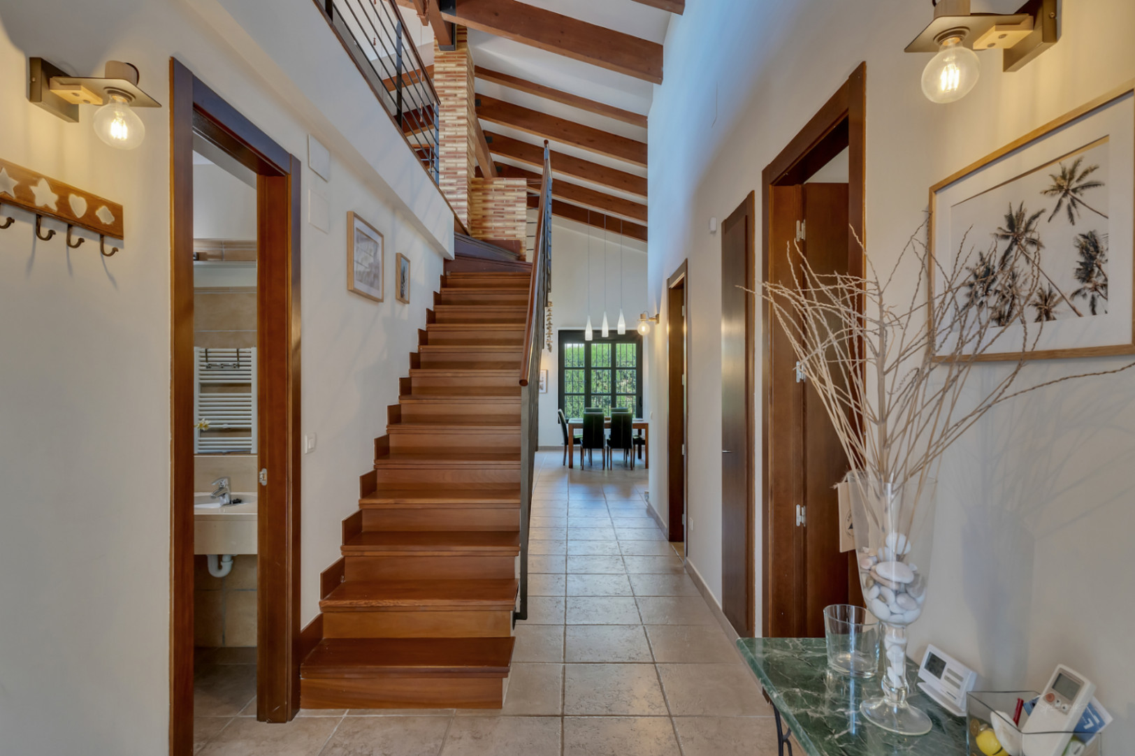 Pasillo distribuidor con techos de doble altura y vigas de madera vistas. Destaca la escalera de madera maciza y el acceso directo al salón-comedor al fondo.