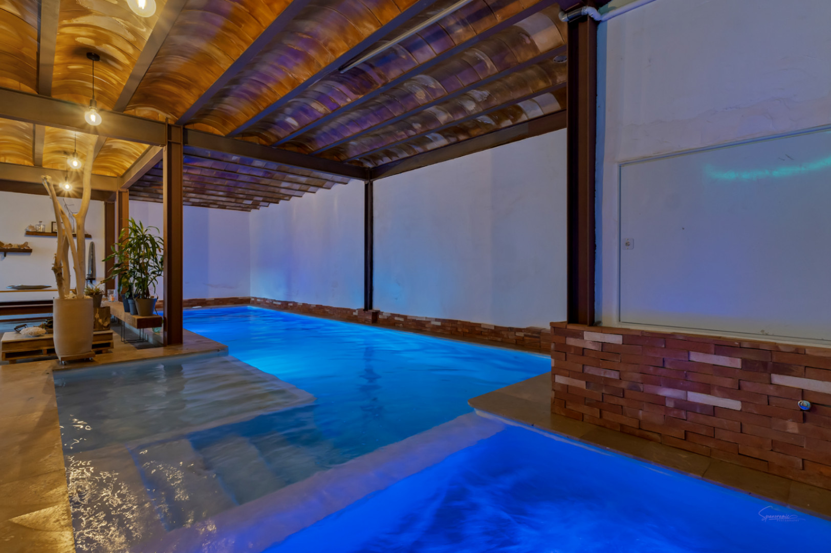Piscina interior climatizada con iluminación LED azul, techos de bovedilla cerámica y vigas de acero. Espacio de spa con jacuzzi integrado y detalles de ladrillo visto.